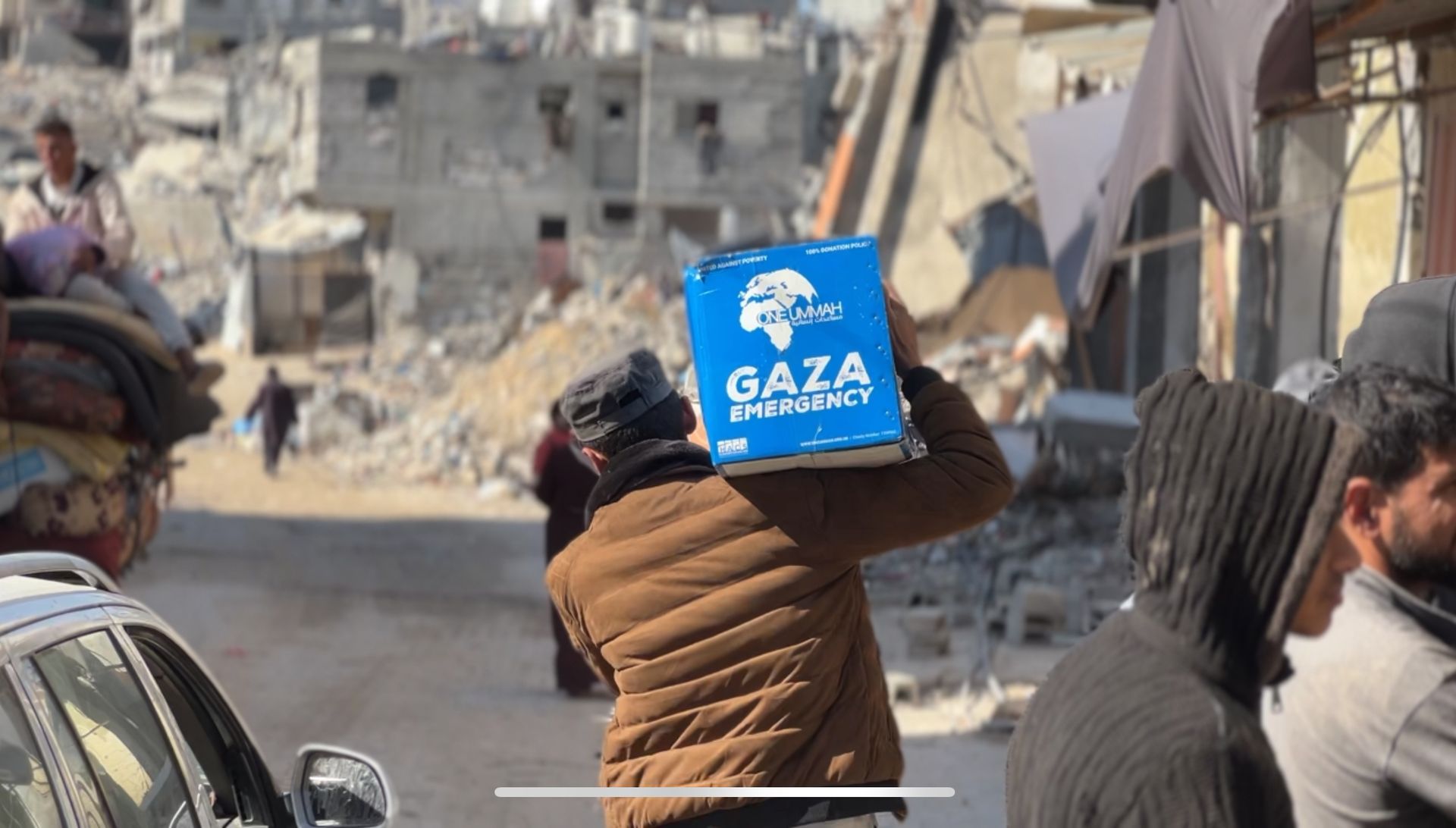 Gaza emergency aid pack on an older man’s back with destruction in the background.